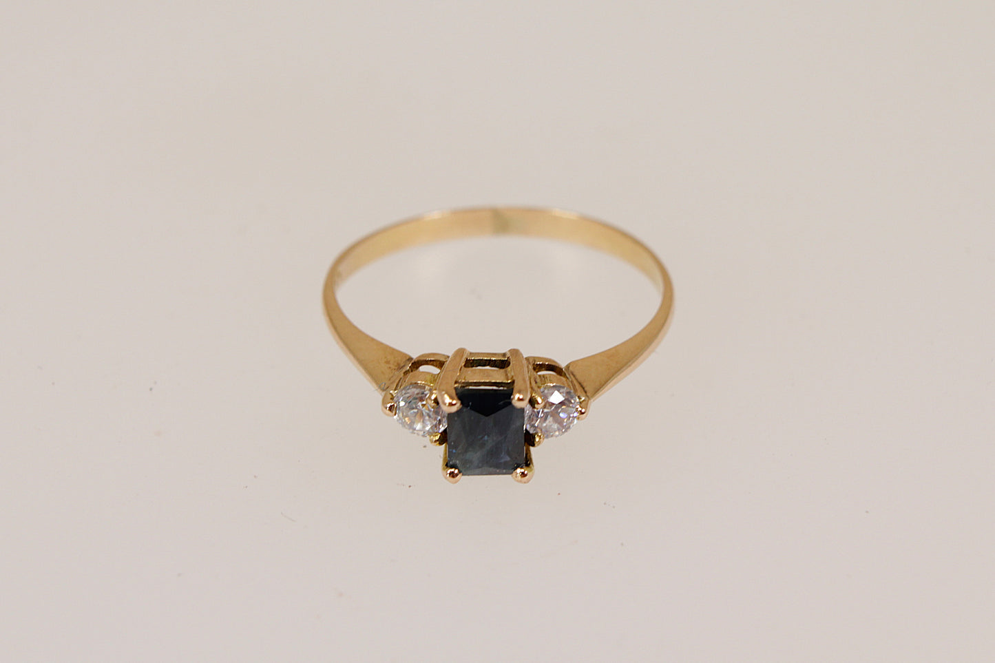 Gold ring with a central blue sapphire  gemstone and two smaller cubic zirconia diamond simulant gemstones  on a light gray background