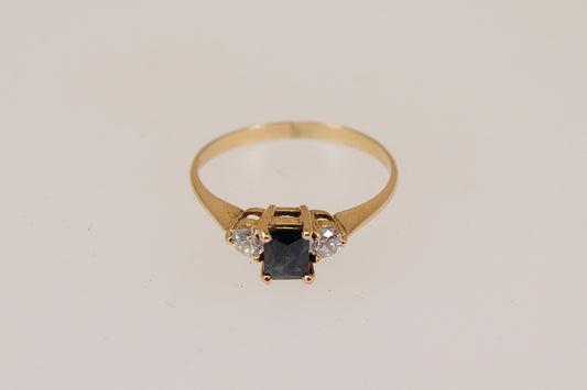 Gold ring with a central blue sapphire  gemstone and two smaller cubic zirconia diamond simulant gemstones  on a light gray background