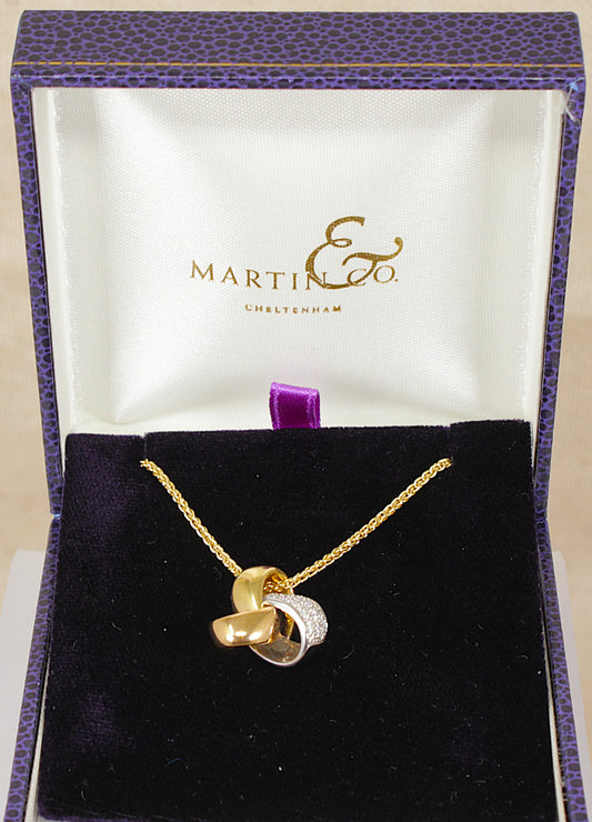 A three tone 18ct gold and diamond  interlocking pendant necklace displayed inside an open Martin & Co. gift box with a purple textured exterior and white satin interior.
