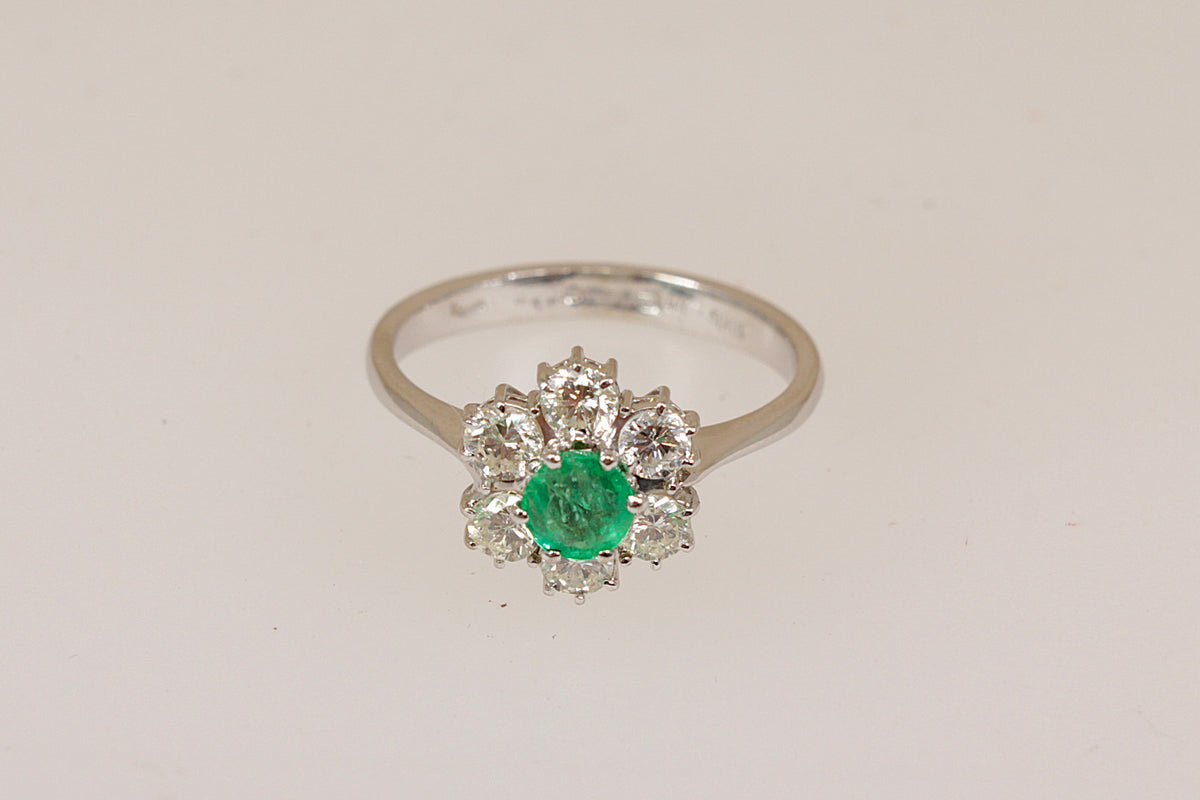 A white 18ct gold ring featuring a central round green emerald gemstone surrounded by a halo of small round diamond gemstones, set on a plain light background. 