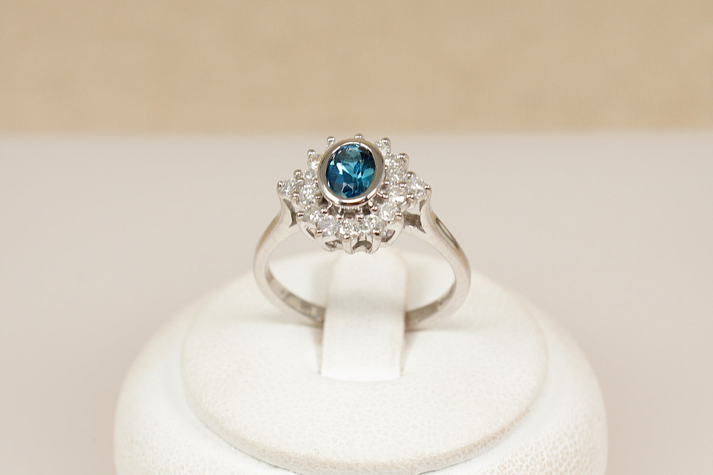 18ct white gold  ring with a blue topaz gemstone surrounded by a halo of  diamonds on a white stand against a beige background