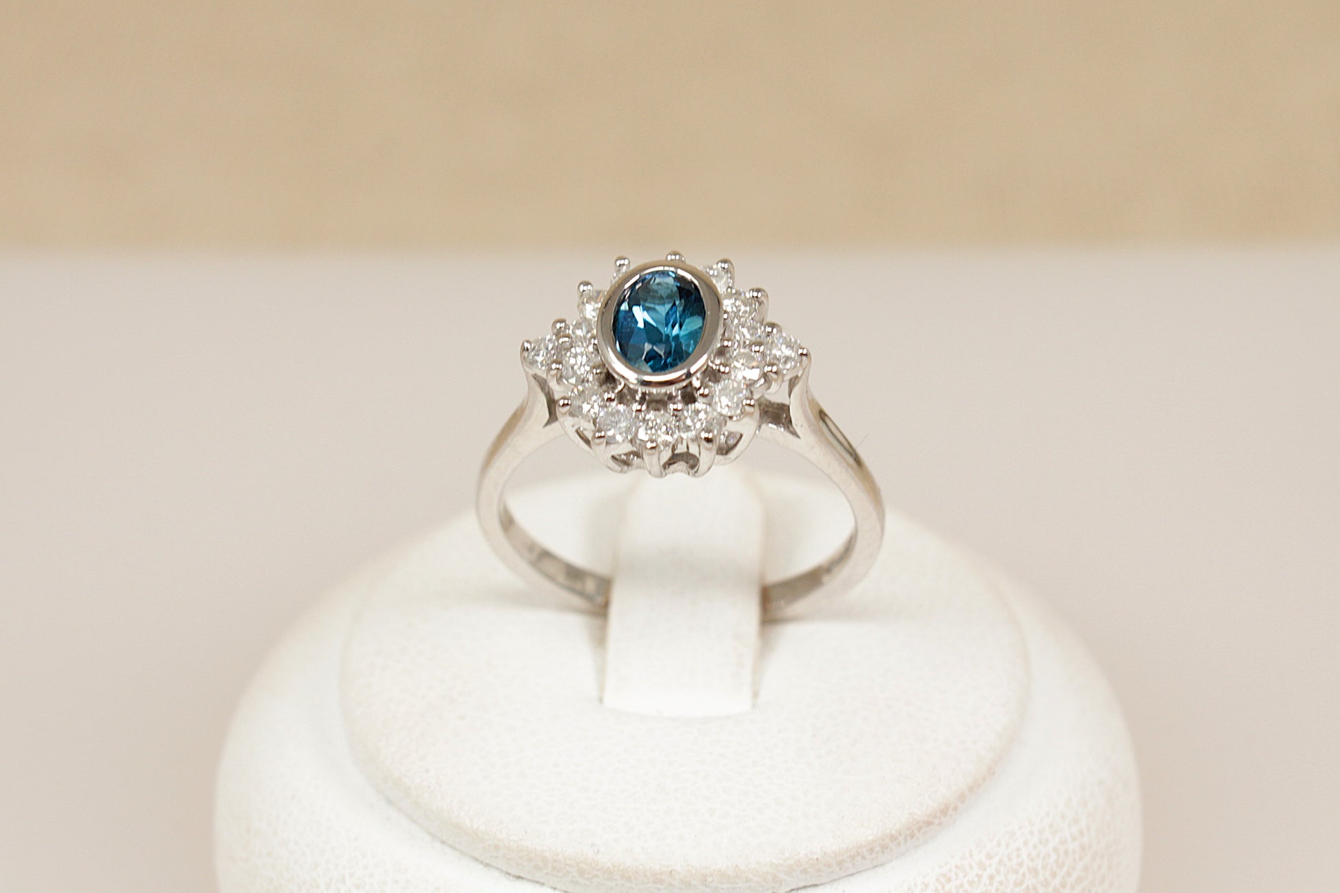 18ct white gold  ring with a blue topaz gemstone surrounded by a halo of  diamonds on a white stand against a beige background