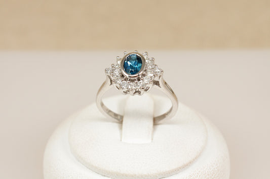 18ct white gold  ring with a blue topaz gemstone surrounded by a halo of  diamonds on a white stand against a beige background