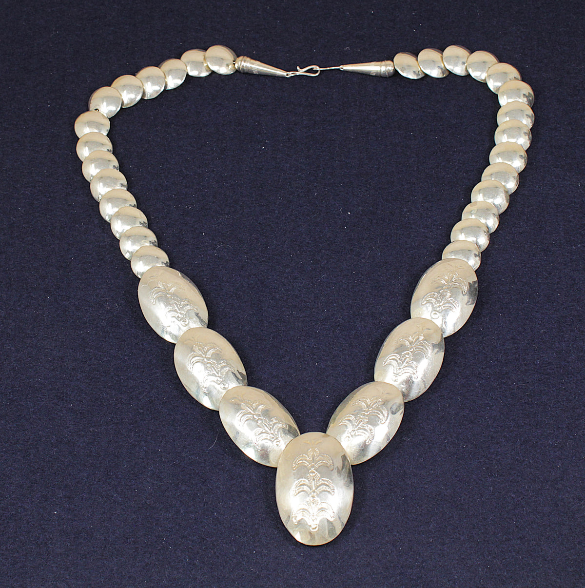A silver-toned statement necklace featuring a strand of small round beads and larger oval pendant links, all engraved with a floral pattern, displayed on a dark navy blue background.