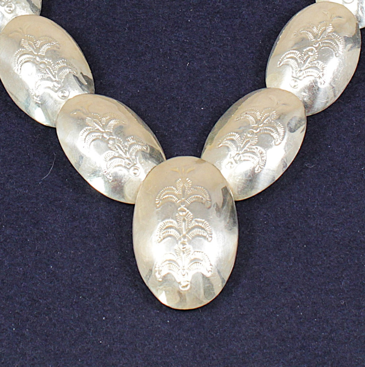 A close-up of a silver-toned necklace with oval links engraved with a floral or scroll motif, displayed against a dark navy background.