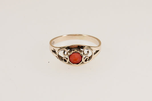 A gold ring with a round coral centerpiece set in a scalloped bezel, with gold heart designs on each shoulder, displayed against a white background.