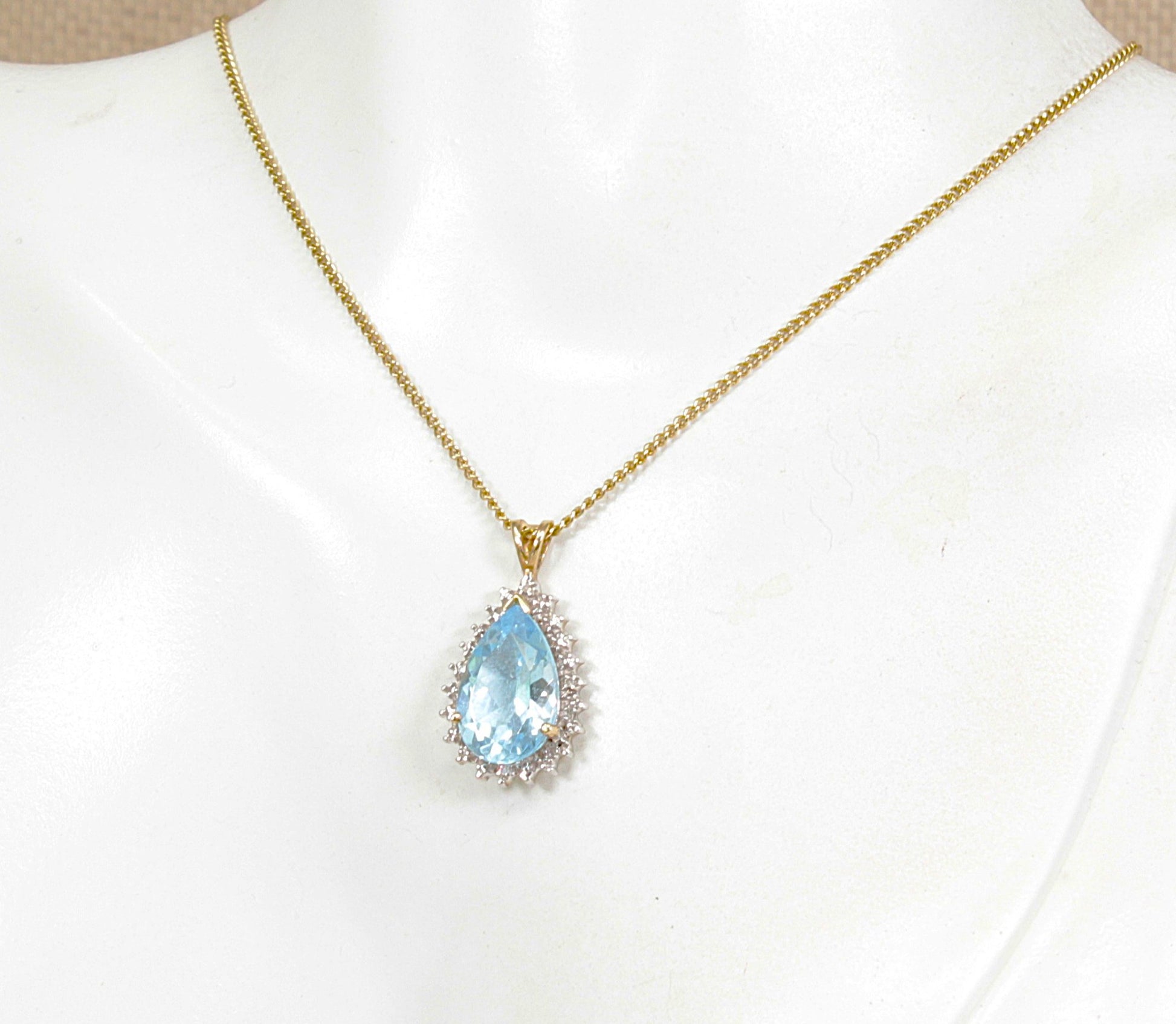 A gold chain necklace with a teardrop-shaped blue topaz gemstone pendant, surrounded by small diamond gemstones, displayed on a white mannequin bust.