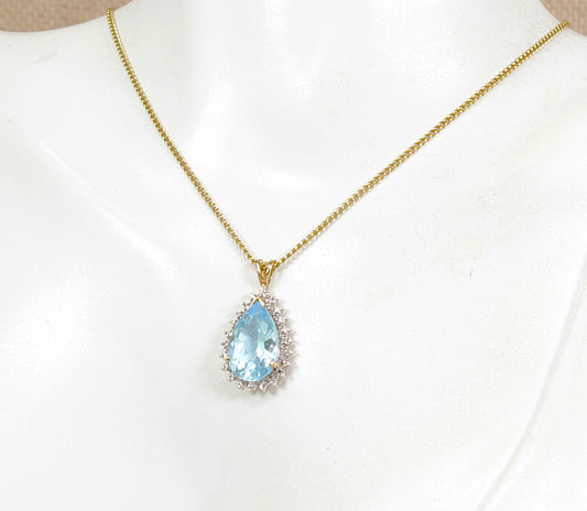 A gold chain necklace with a teardrop-shaped blue topaz gemstone pendant, surrounded by small diamond gemstones, displayed on a white mannequin bust.
