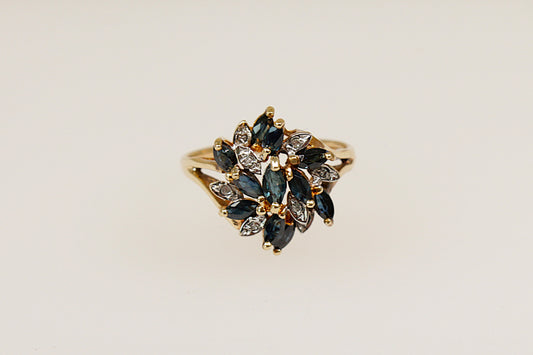 This 9ct Gold ring is a breathtaking and artistic statement piece! This large Sapphire and Diamond flower design ring features 11 Marquise-cut blue Sapphires in claw settings and 8 round diamond accents, Birmingham hallmarks for year 1994 and stamped DIA. UK finger size O. 