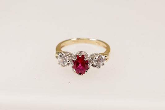 A 9ct gold trilogy ring with an oval cut ruby centerpiece and two round cut crystal gemstones on either side, set in a crown-inspired setting.