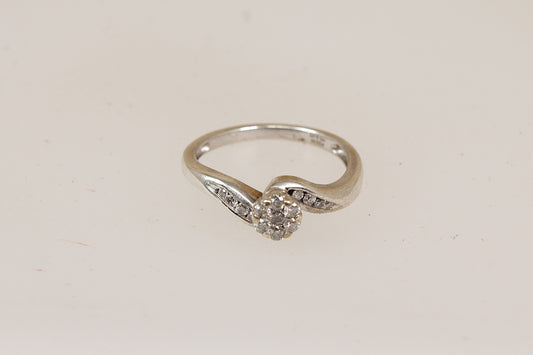 A silver ring with a curved band and a central round-cut diamond gemstones, accented by small diamonds along the shoulders, shown on a plain light background.