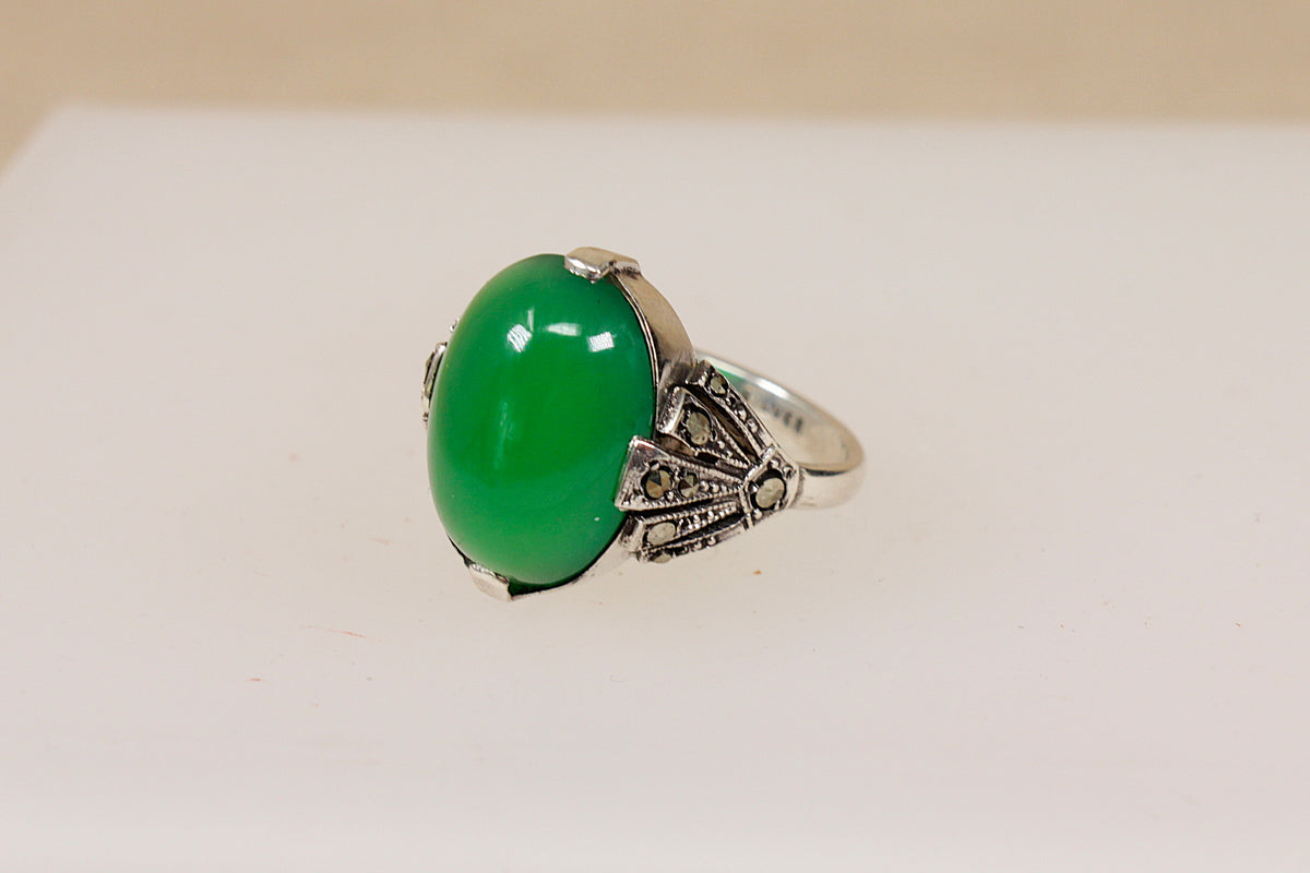 A silver ring featuring a large, smooth oval green chrysoprase gemstone, set in an ornate Art Deco style band with small  marcasite gemstones, shown on a plain light background.