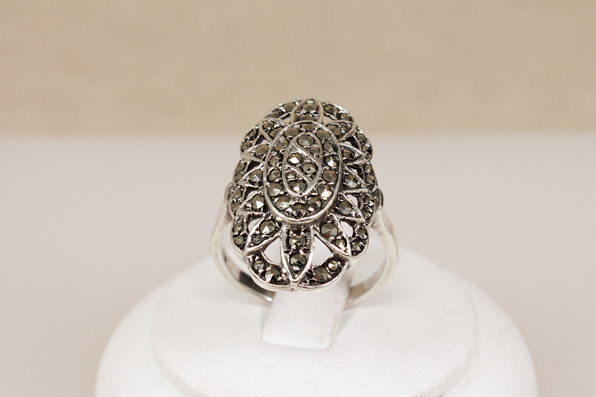 An ornate art deco silver ring with a large oval filigree design set with small  marcasite gemstones, displayed on a white stand against a soft beige background.