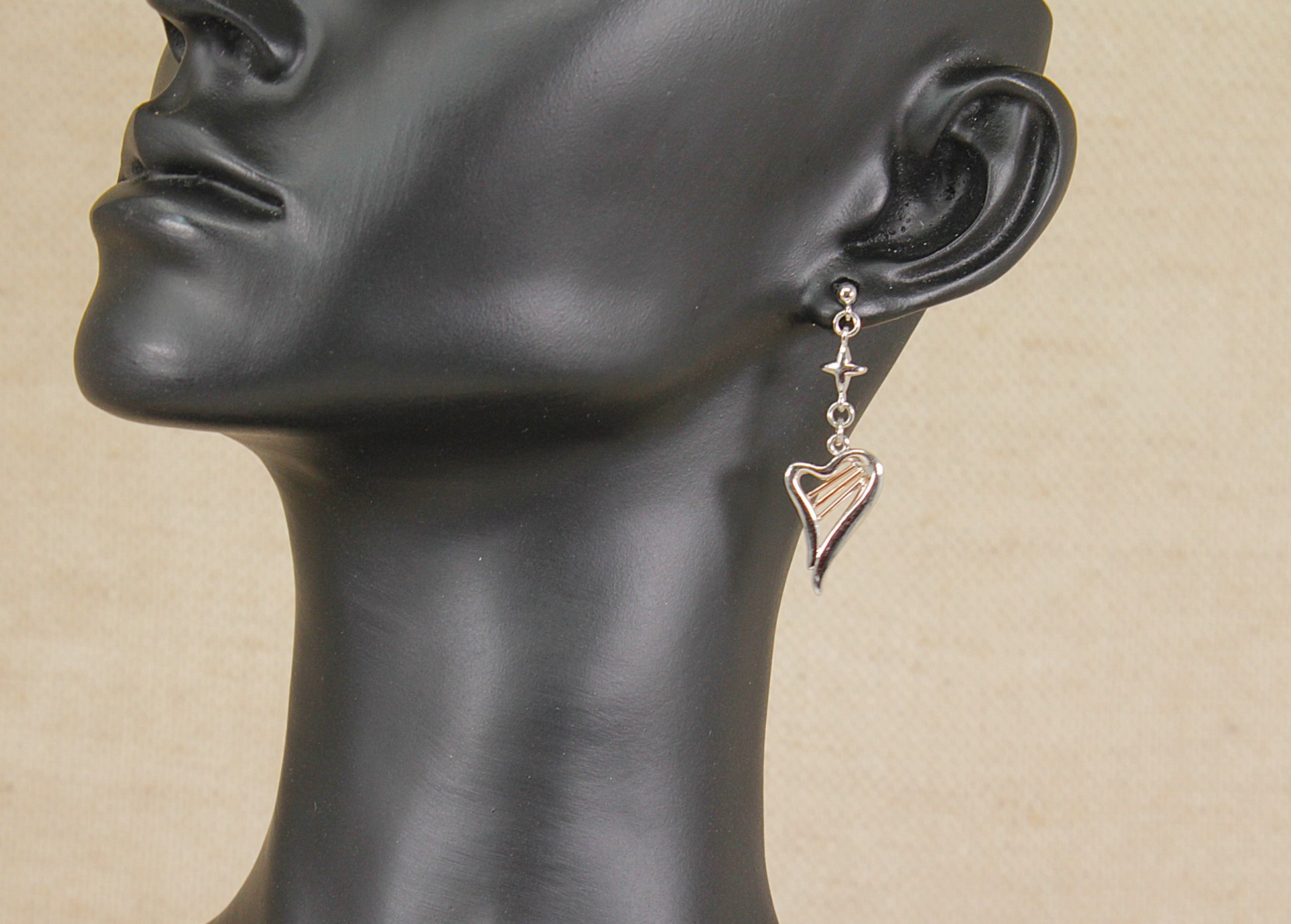 A Clogau silver and gold dangle earring in the shape of a harp with a small cross  charm, shown on a black mannequin head against a beige background.