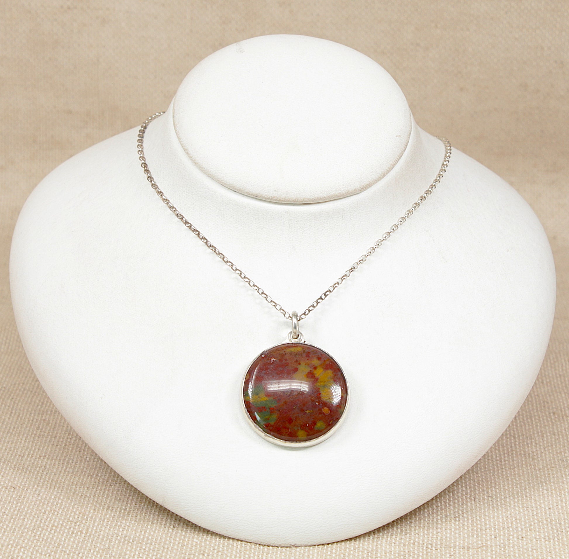 A silver chain necklace with a round cabochon pendant featuring an mottled brown, and green gemstone, displayed on a white jewelry bust against a beige fabric background.