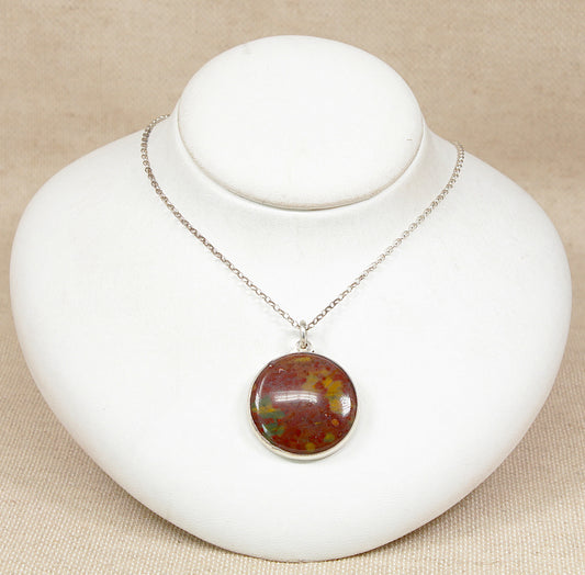 A silver chain necklace with a round cabochon pendant featuring an mottled brown, and green gemstone, displayed on a white jewelry bust against a beige fabric background.