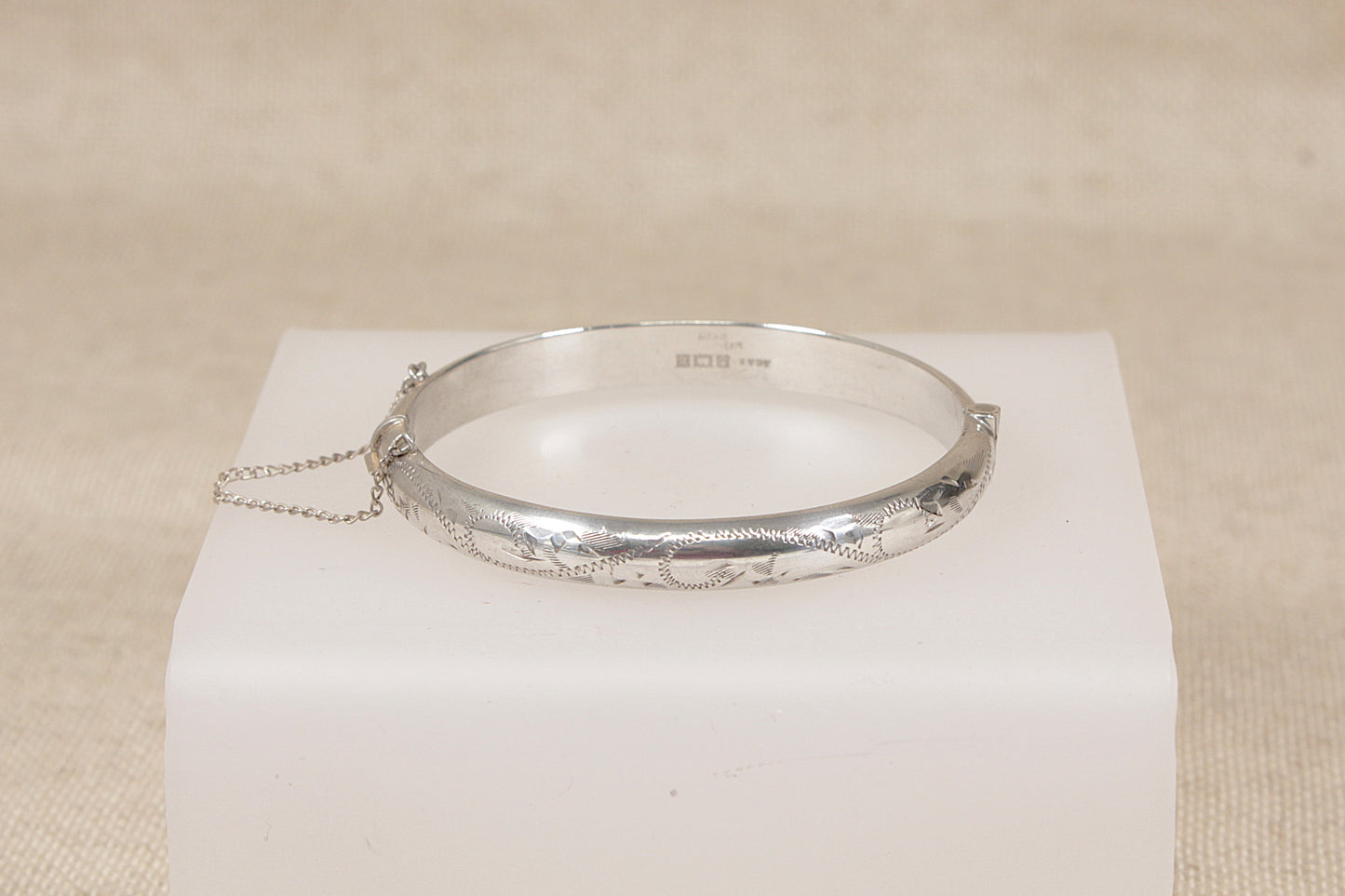 A vintage silver hinged bangle bracelet with engraved floral details, resting on a white display block against a neutral beige background.