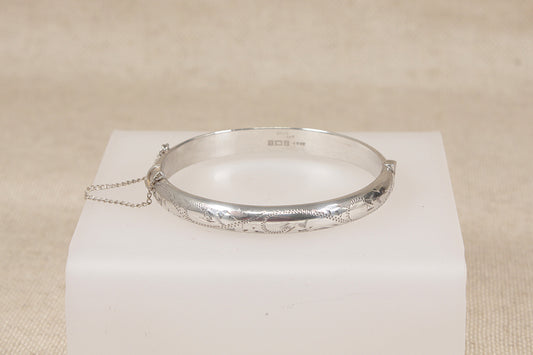 A vintage silver hinged bangle bracelet with engraved floral details, resting on a white display block against a neutral beige background.