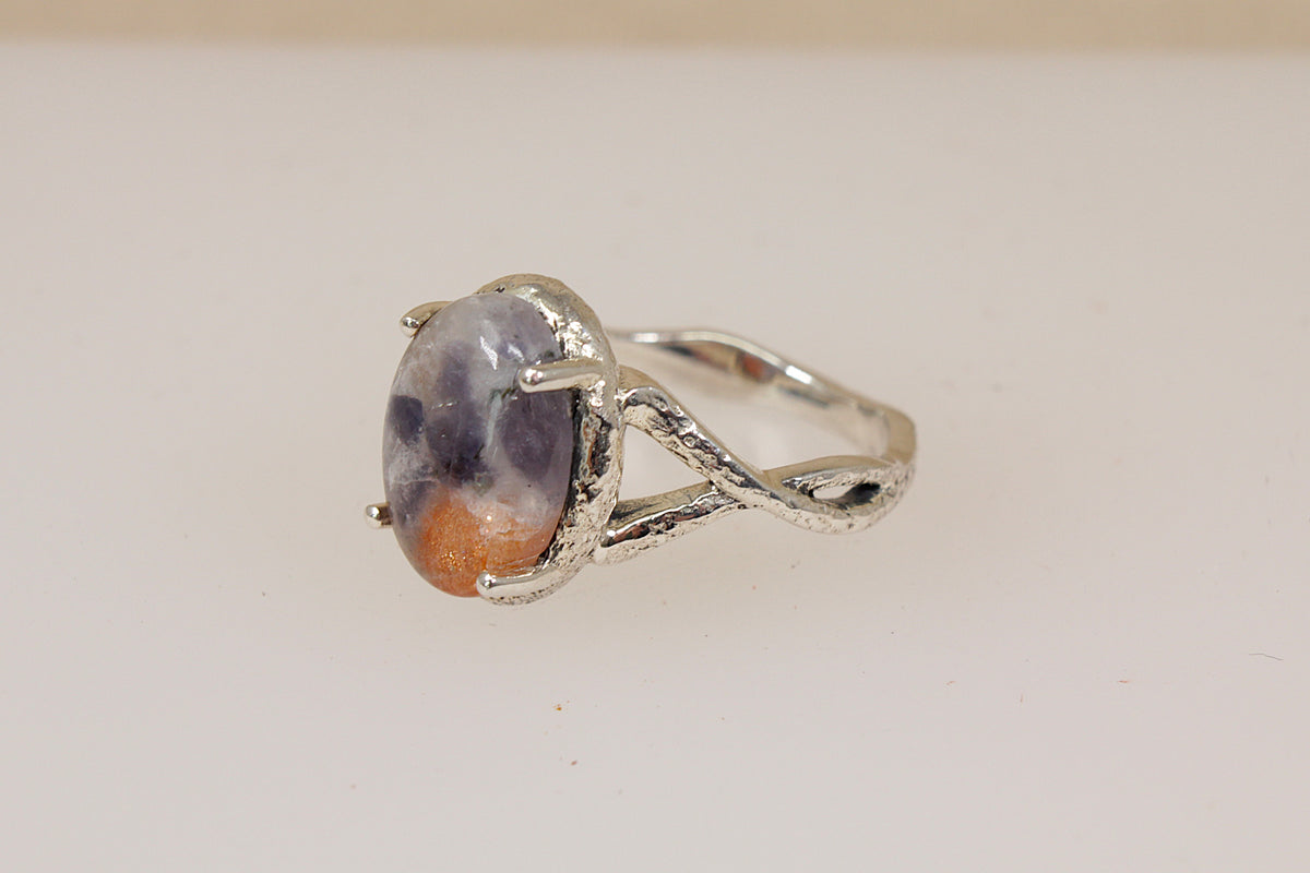 A silver ring featuring an oval cabochon gemstone with a marbled pattern of purple, gray, and orange-brown, set in a branch-like prong design on a plain light background.