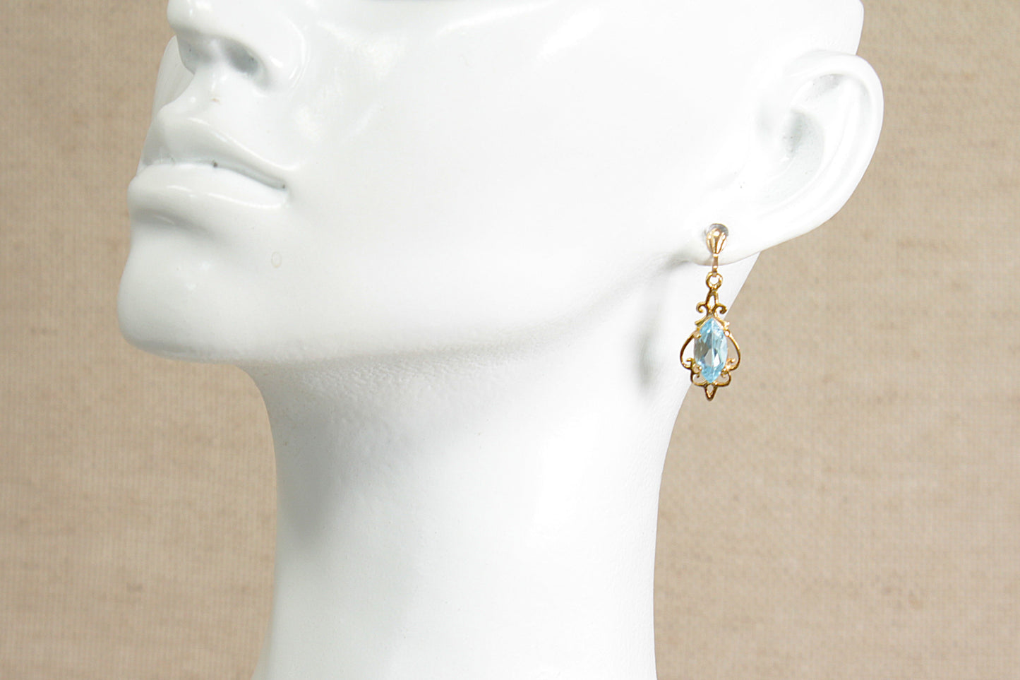 A gold dangle earring with a blue topaz teardrop-shaped gemstone, displayed on a white ceramic mannequin head against a neutral beige background.