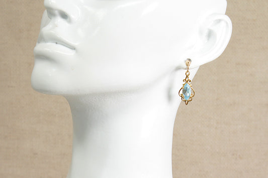 A gold dangle earring with a blue topaz teardrop-shaped gemstone, displayed on a white ceramic mannequin head against a neutral beige background.