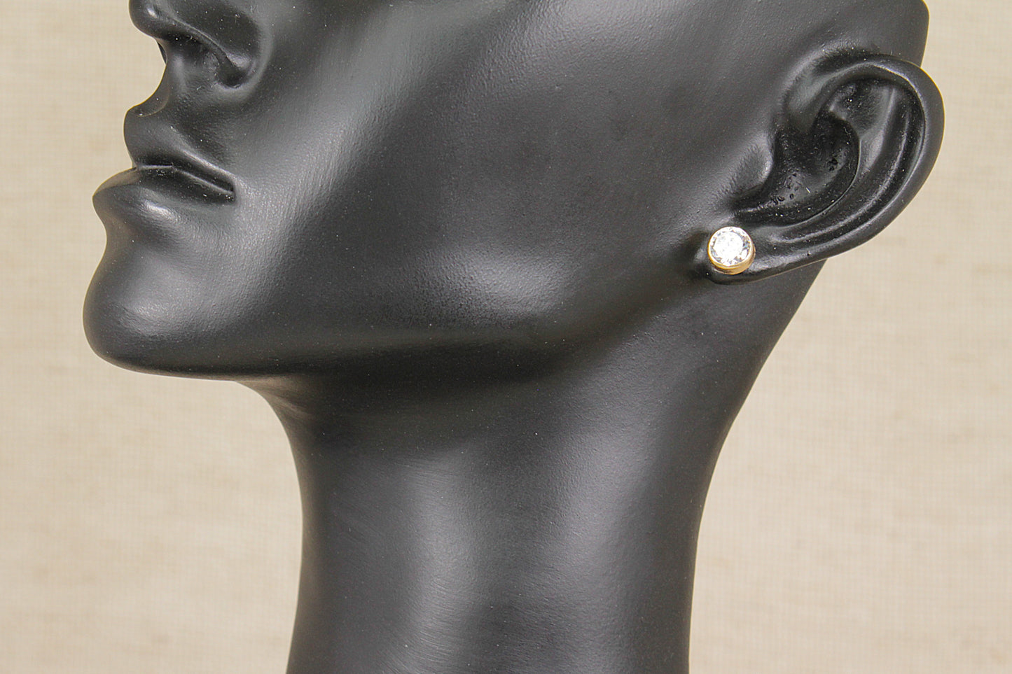 A close-up side profile of a black mannequin head wearing a small gold stud earring with a clear round gemstone, set against a light beige background.