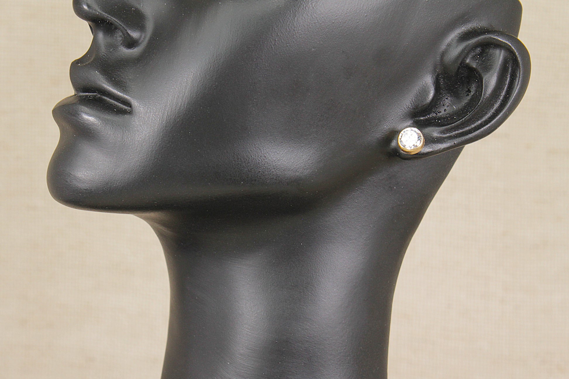 A close-up side profile of a black mannequin head wearing a small gold stud earring with a clear round gemstone, set against a light beige background.