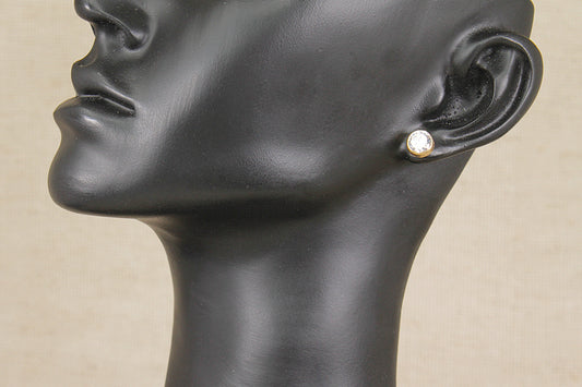 A close-up side profile of a black mannequin head wearing a small gold stud earring with a clear round gemstone, set against a light beige background.