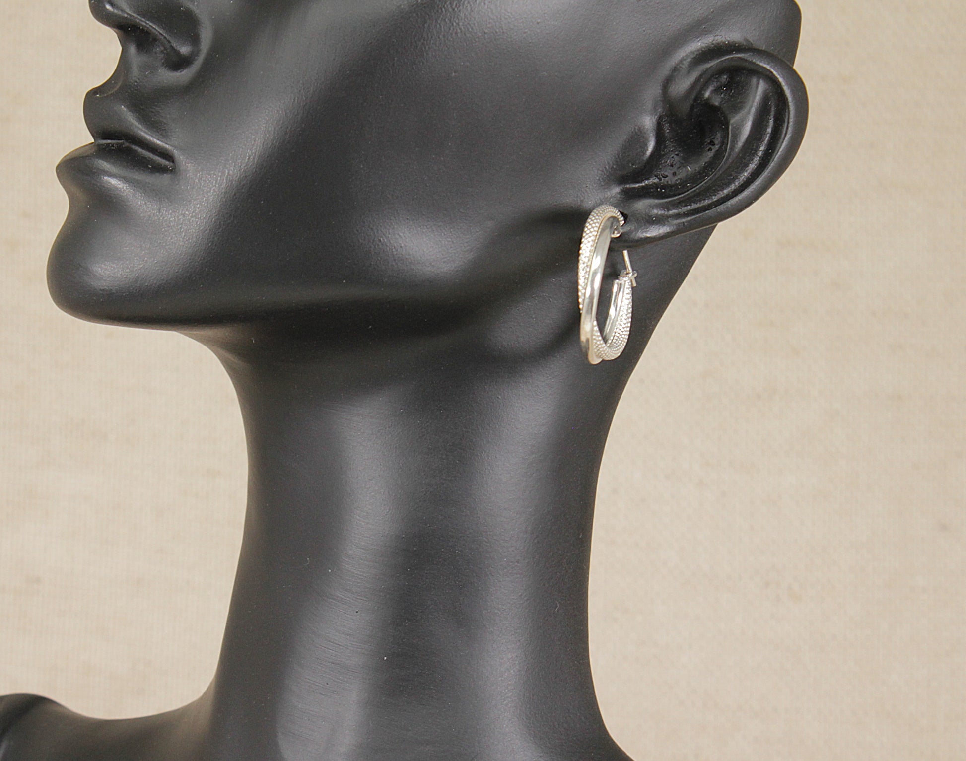 A silver textured hoop earring worn on the ear of a black mannequin head, shown in a close-up side profile against a neutral beige background.
