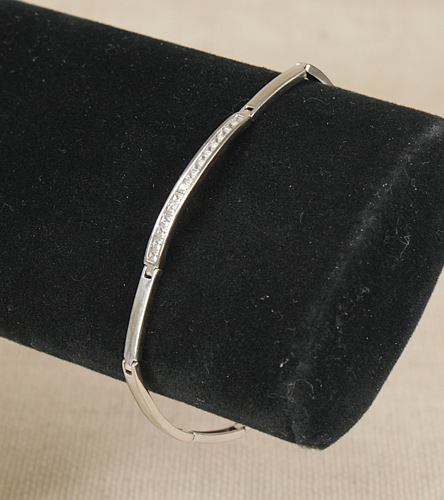 A sterling silver bracelet with a row of small clear white crystal  gemstones along one side, displayed on a black velvet jewelry stand against a light beige background. 