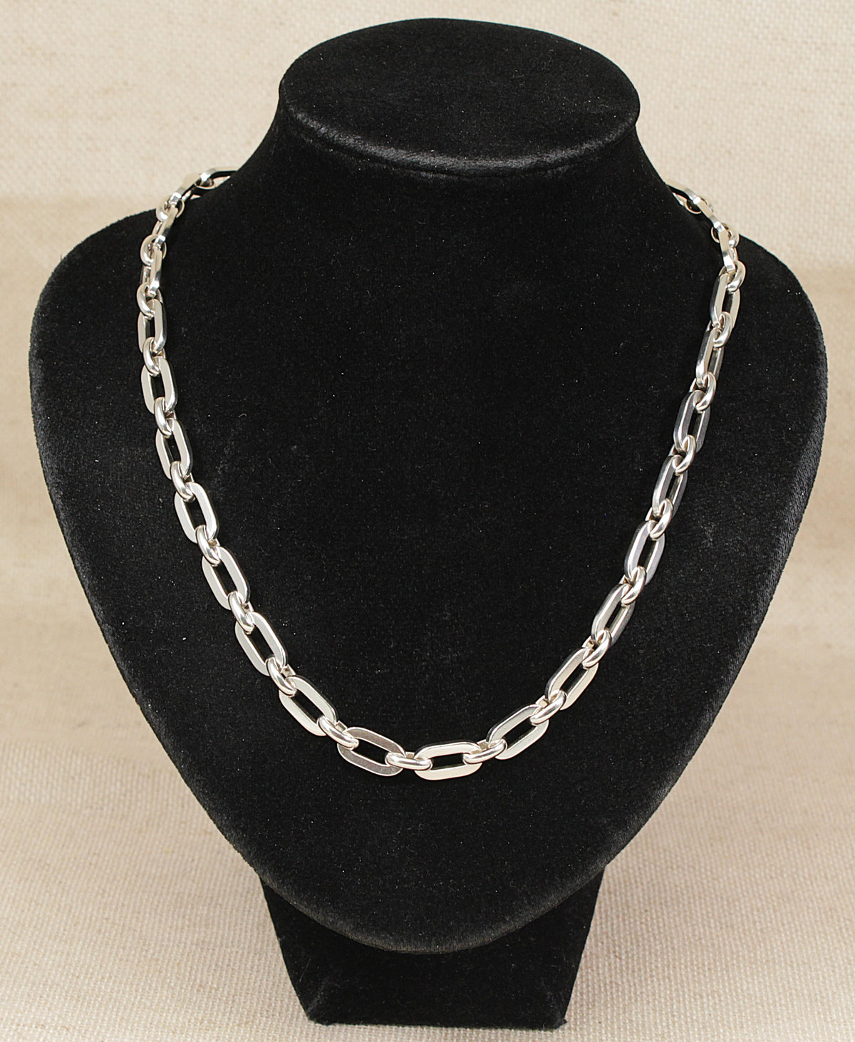 A silver-toned chain necklace with elongated oval links, displayed on a black velvet jewelry bust against a light beige textured background.
