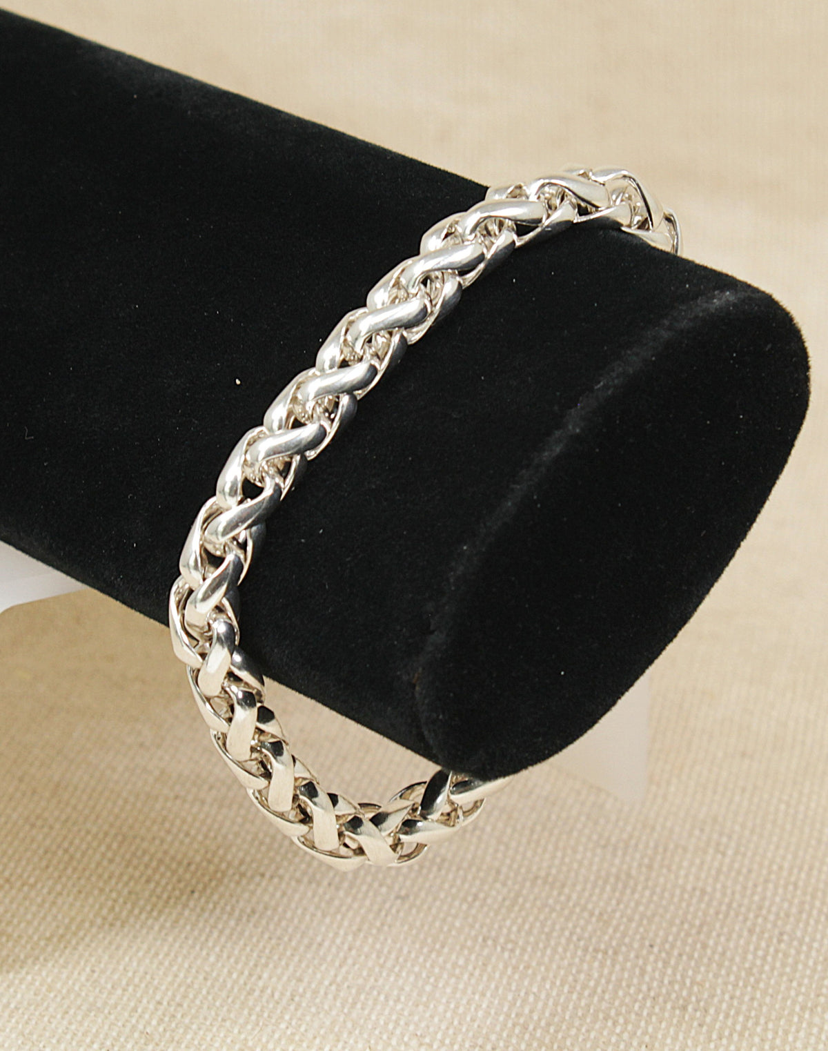 A silver-toned rope chain bracelet displayed on a black cylindrical velvet jewelry stand against a light beige fabric background.