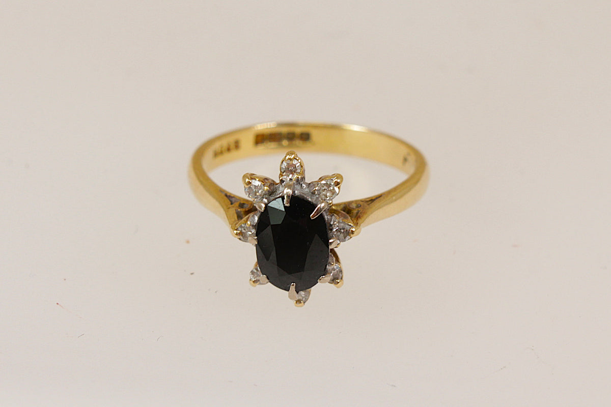 A yellow gold ring featuring an oval-cut midnight blue sapphire gemstone in a prong setting, accented by small diamond  gemstones, shown on a plain light background.