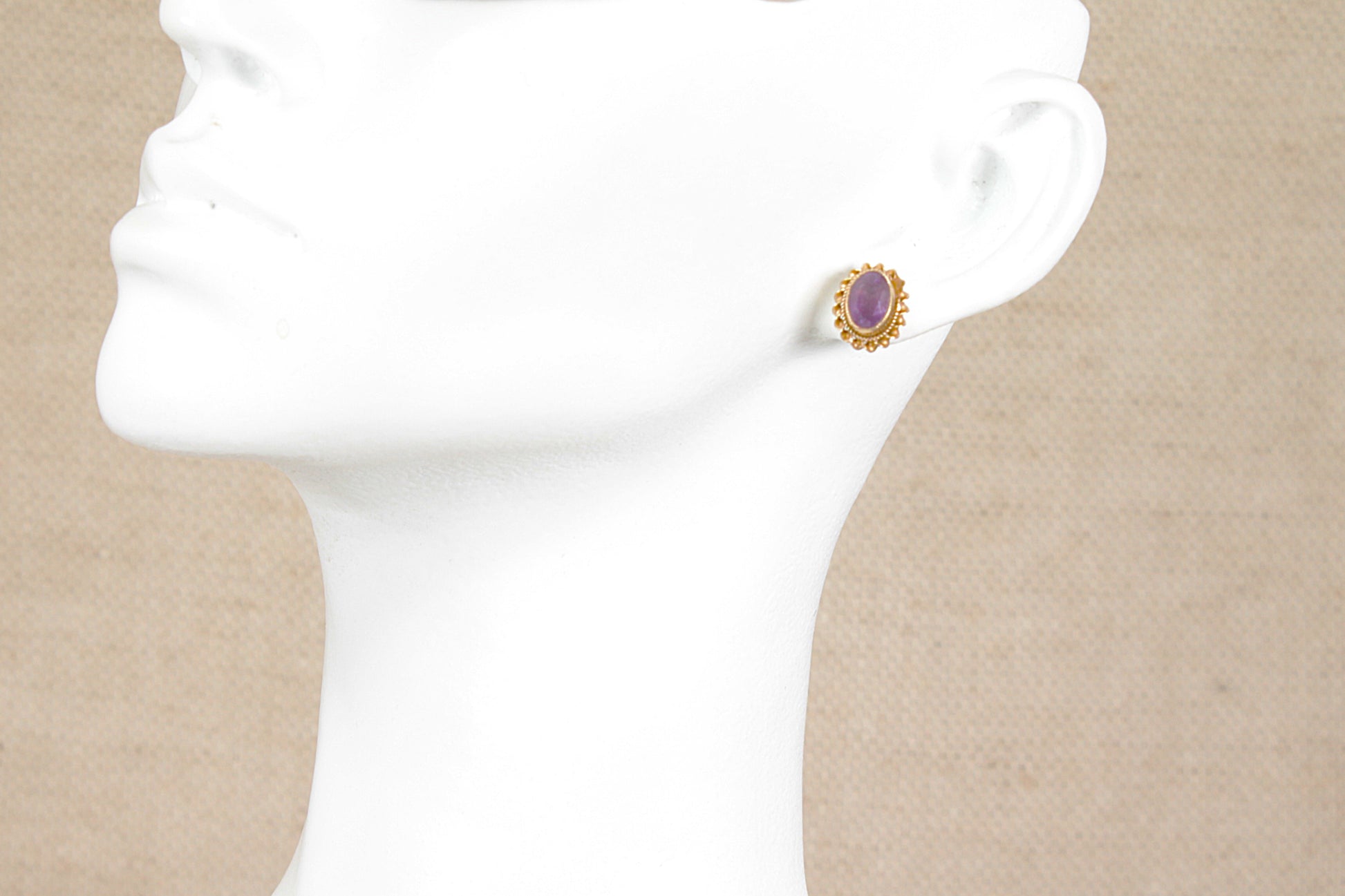 A single 9ct gold stud earring with an oval purple amethyst gemstone, displayed on a white mannequin head against a beige textured background.