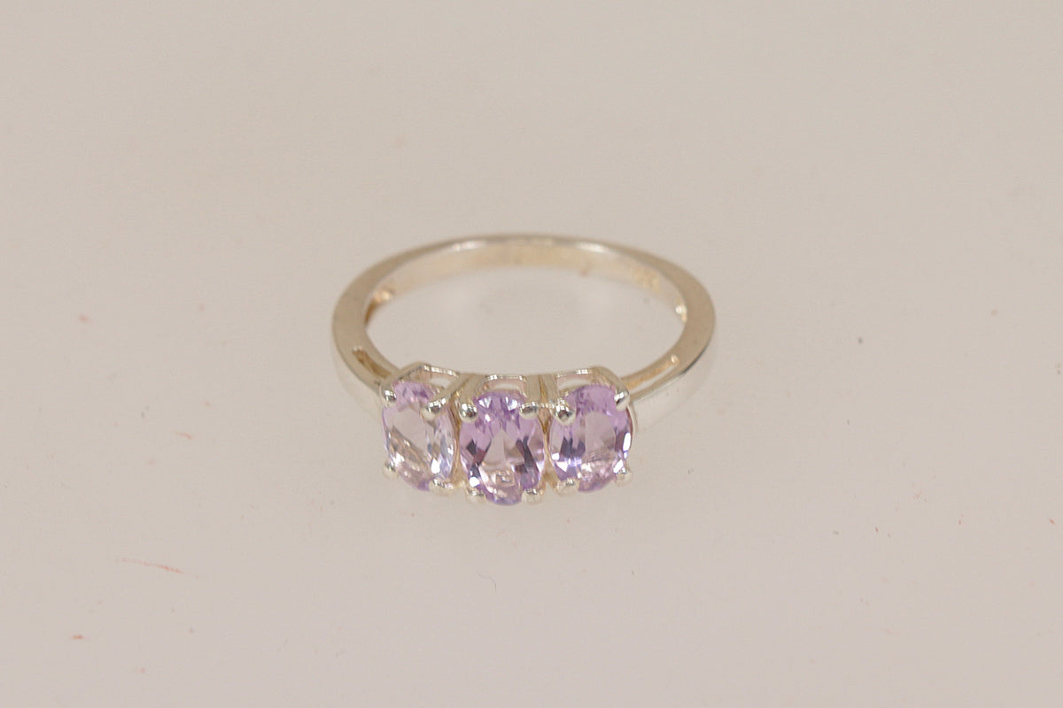 A silver three-stone ring featuring oval-cut light purple rose de france amethyst gemstones, set against a plain off-white background.