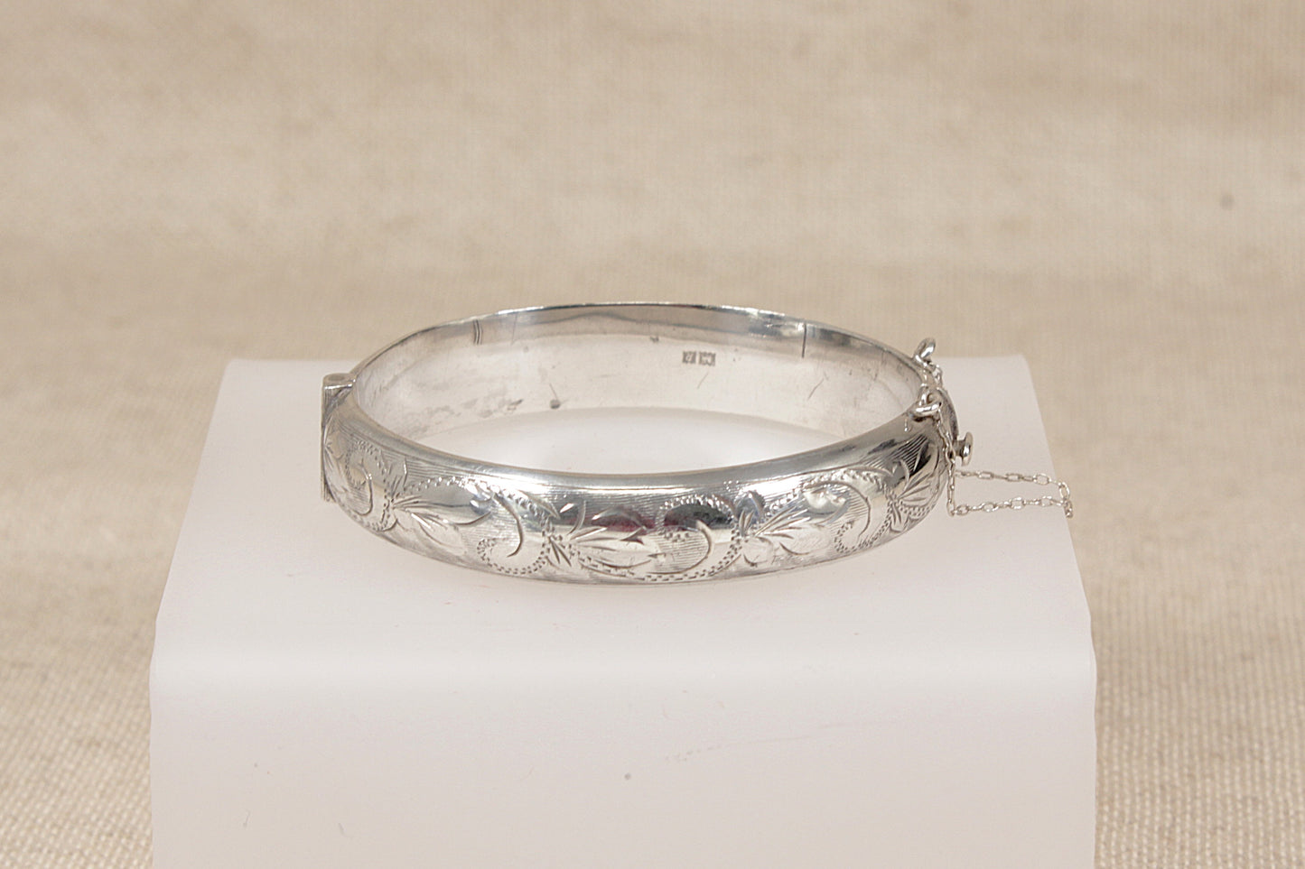 A vintage silver bangle bracelet with engraved floral and scrollwork patterns, displayed on a white rectangular block against a neutral beige fabric background.