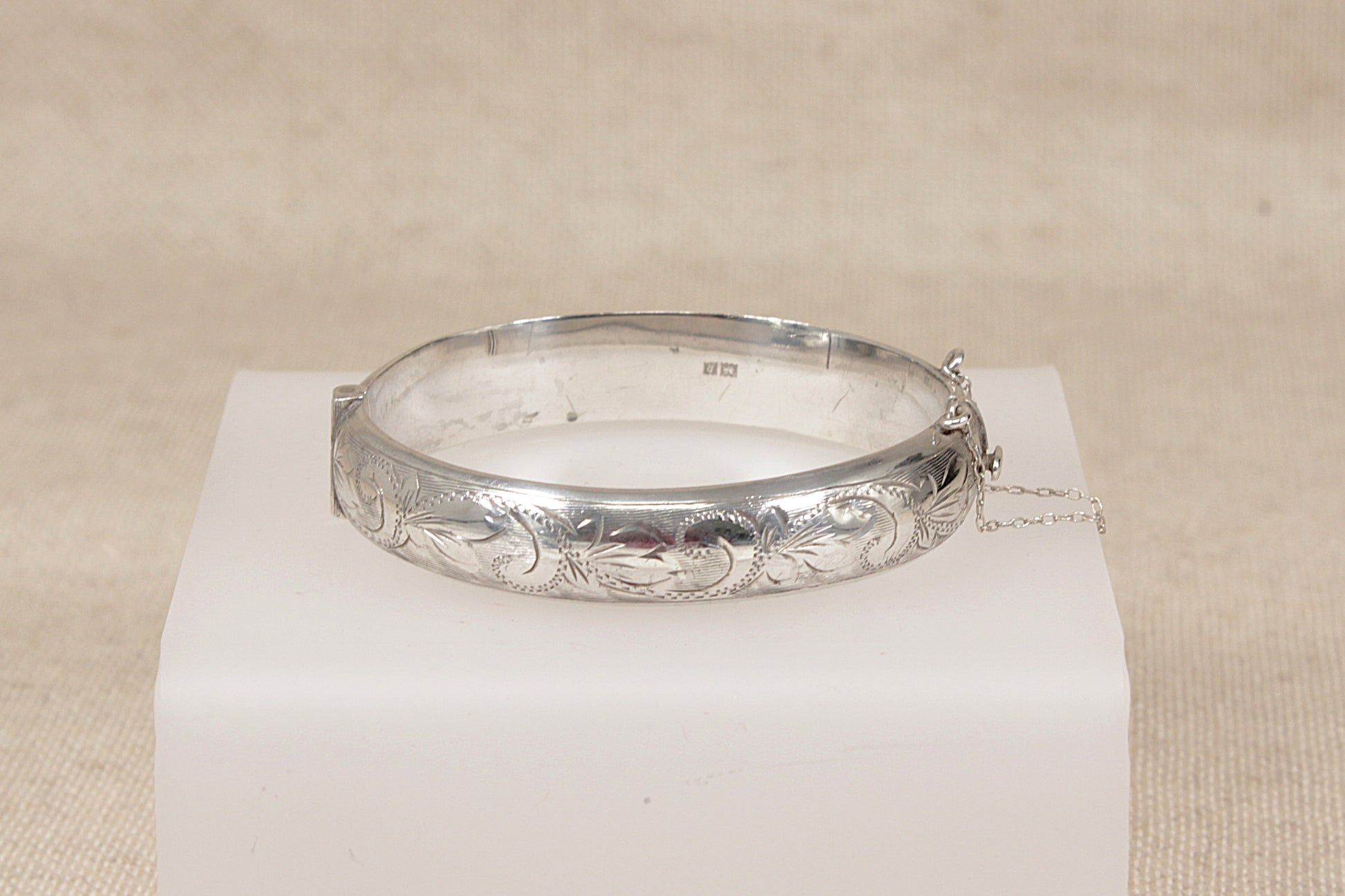 A vintage silver bangle bracelet with engraved floral and scrollwork patterns, displayed on a white rectangular block against a neutral beige fabric background.