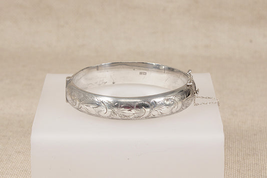 A vintage silver bangle bracelet with engraved floral and scrollwork patterns, displayed on a white rectangular block against a neutral beige fabric background.