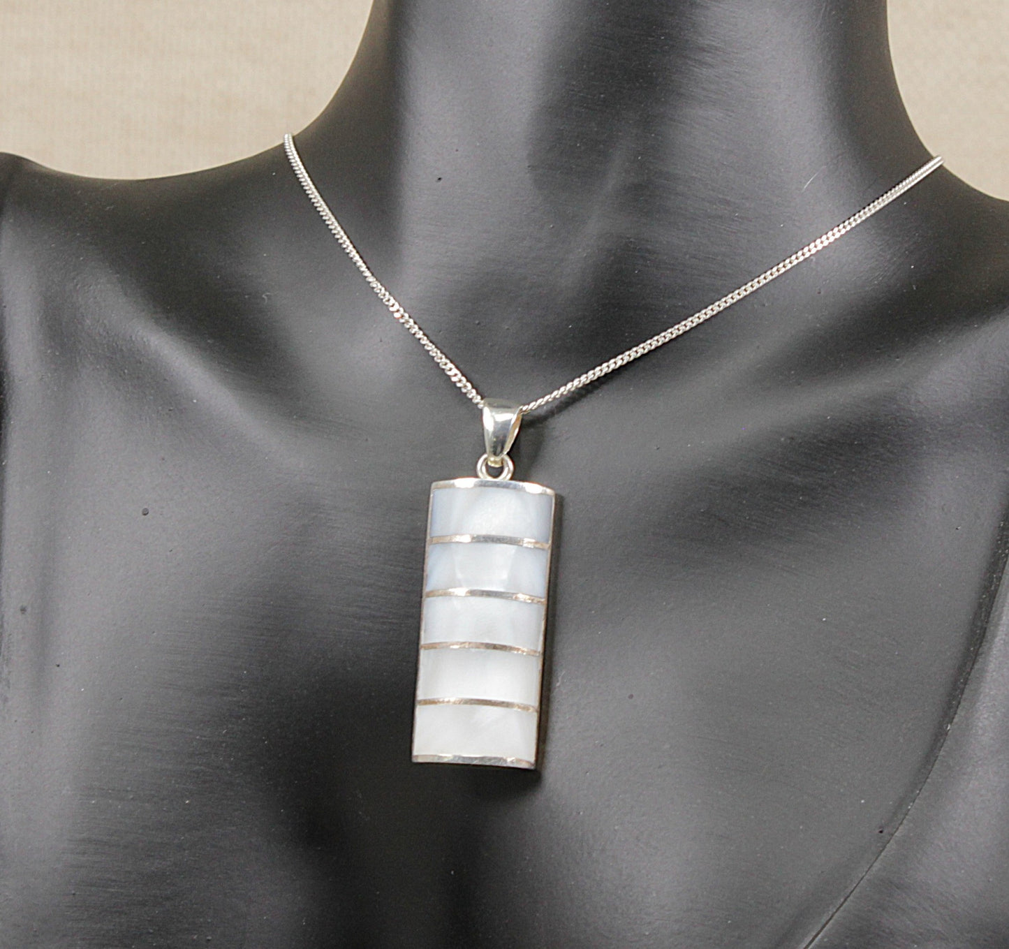 A silver chain necklace with a rectangular pendant featuring horizontal bands of white mother-of-pearl, displayed on a black mannequin bust.