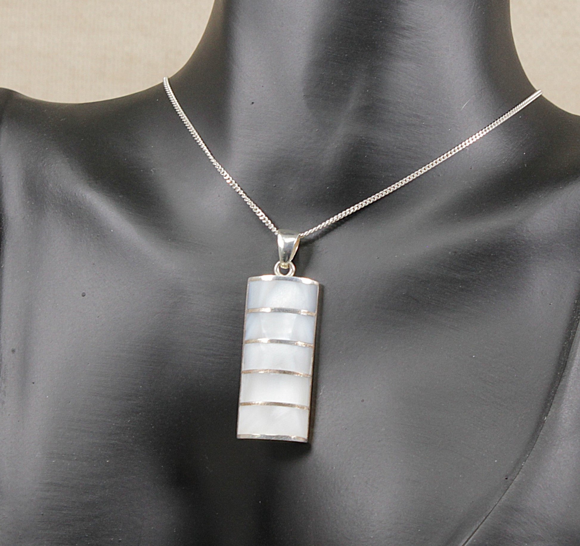 A silver chain necklace with a rectangular pendant featuring horizontal bands of white mother-of-pearl, displayed on a black mannequin bust.