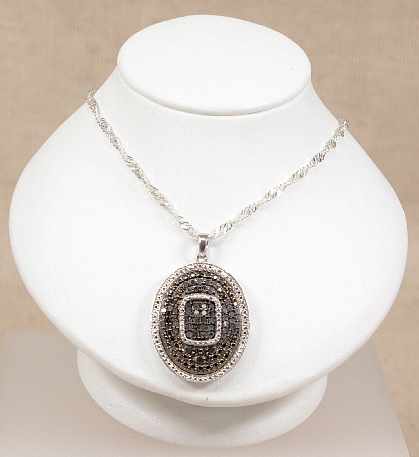 A sterling silver chain necklace with an oval sapphire pendant featuring a square inset, displayed on a white bust  with a beige background.