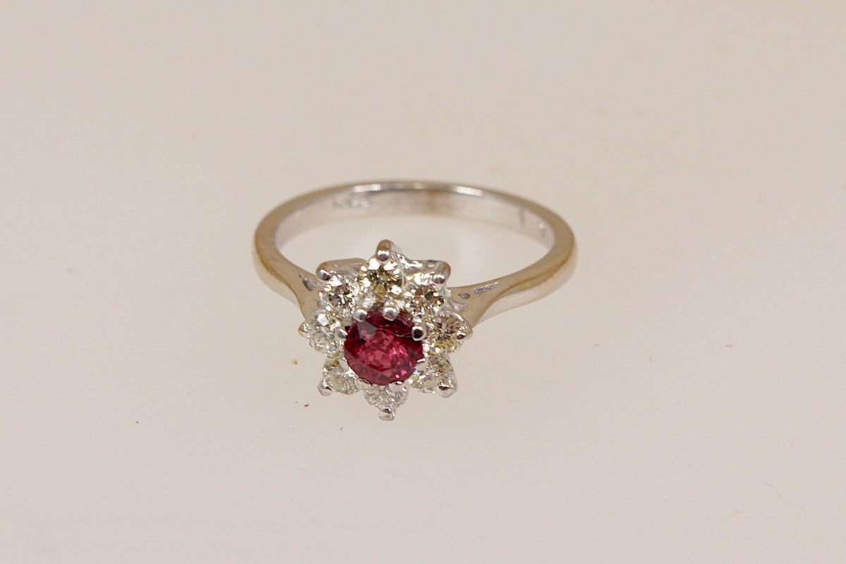 An 18ct white gold ring featuring a round red ruby center gemstone surrounded by a halo of small diamond gemstones, set on a plain light beige background.