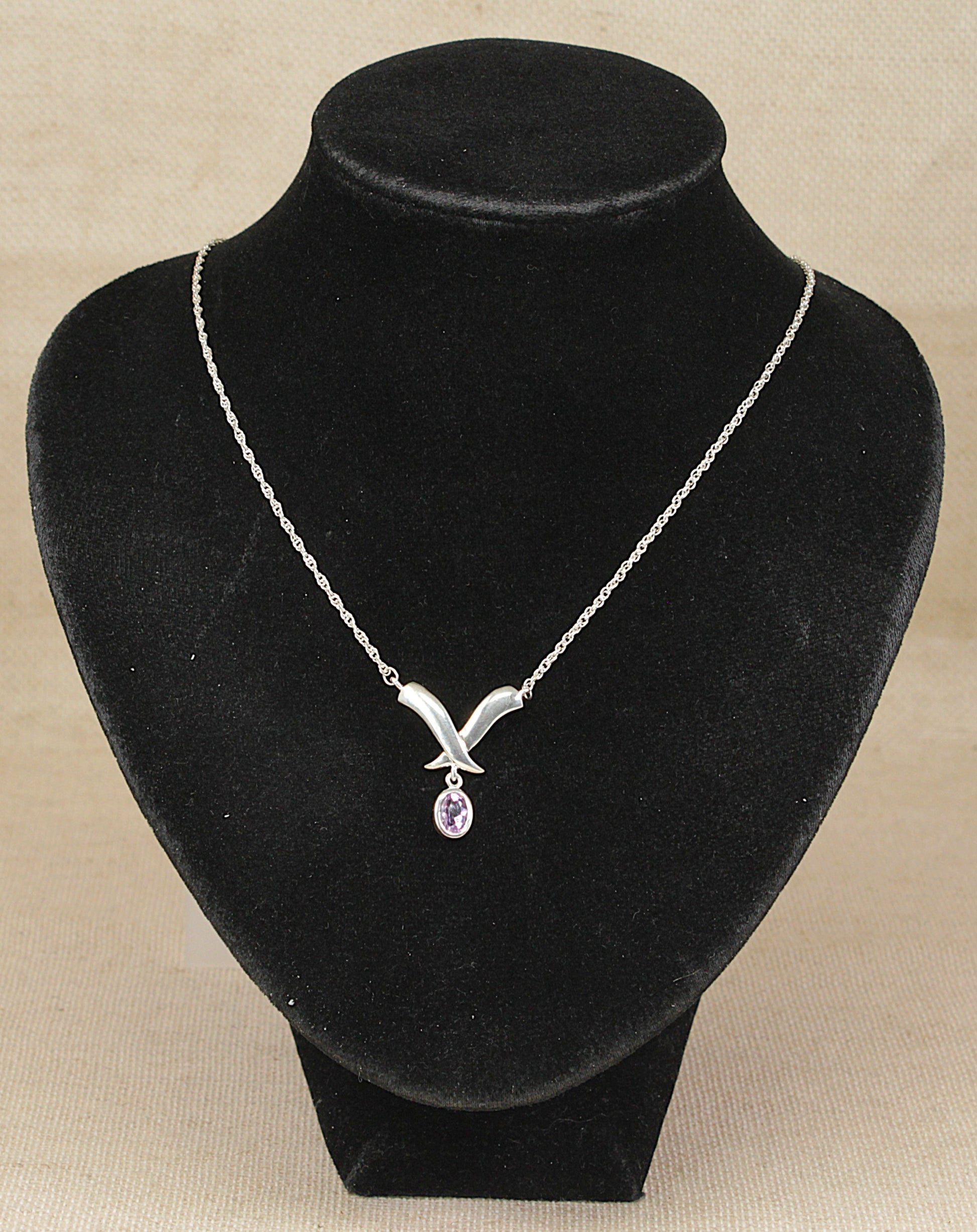 A silver necklace with a stylized V-shaped pendant and a small teardrop purple amethyst gemstone, displayed on a black velvet jewelry bust against a beige background.