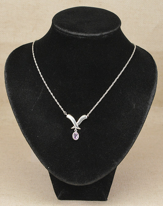 A silver necklace with a stylized V-shaped pendant and a small teardrop purple amethyst gemstone, displayed on a black velvet jewelry bust against a beige background.