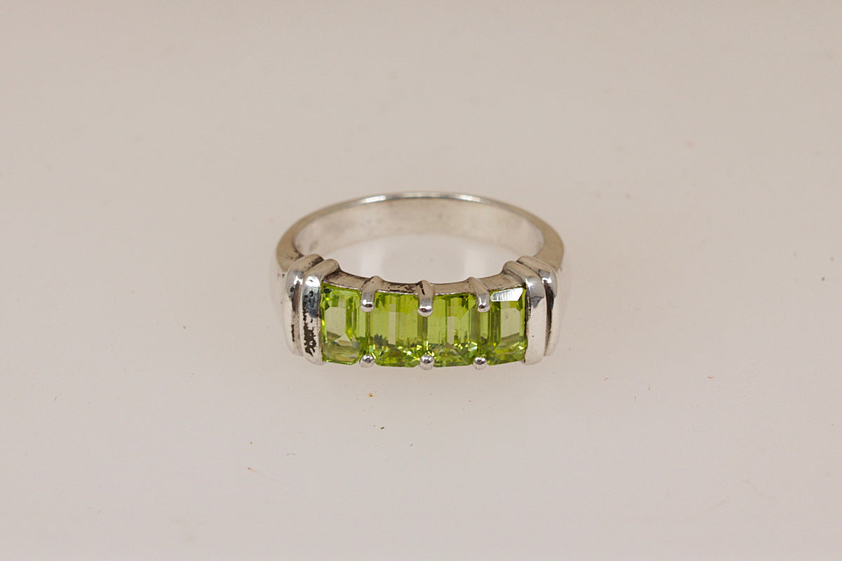 A silver ring featuring a row of rectangular green peridot gemstones, set in a polished silver band, shown on a plain light beige background.