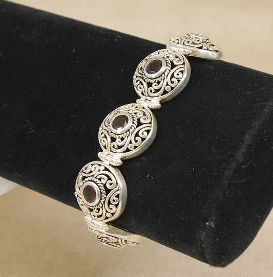 A silver and garnet filigree bracelet displayed on a black cylindrical jewelry stand against a beige fabric background.