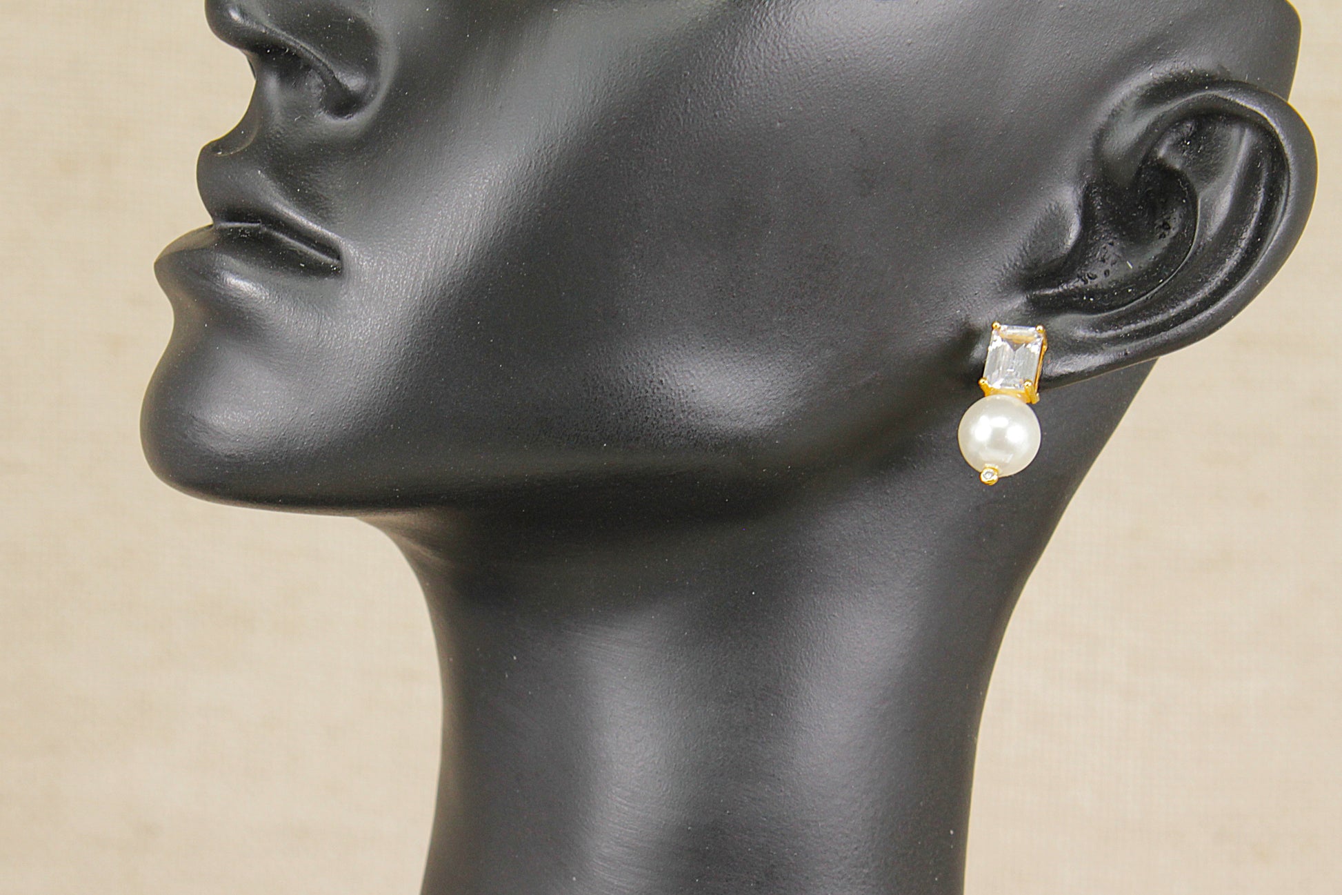 A close-up side profile of a black mannequin head wearing a single drop earring with a small gold-toned top and a white pearl below, against a light beige background.