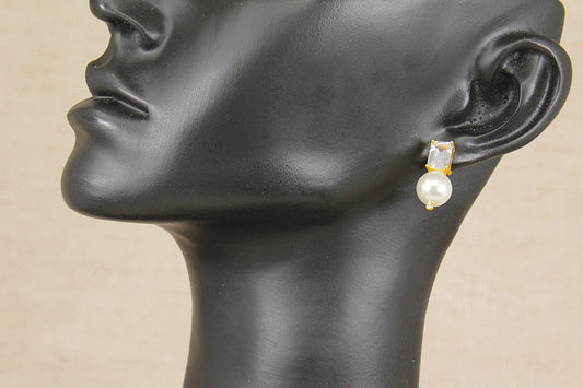 A close-up side profile of a black mannequin head wearing a single drop earring with a small gold-toned top and a white pearl below, against a light beige background.