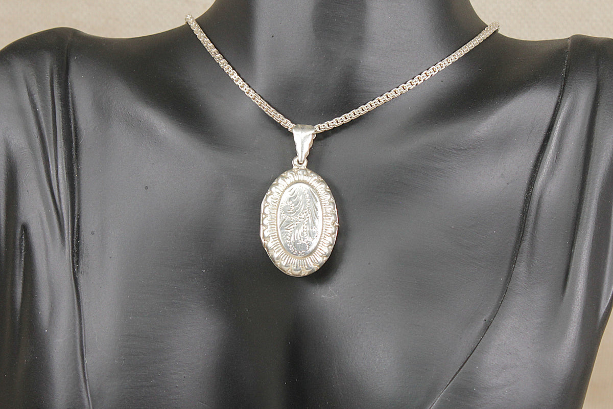 A silver oval pendant necklace with a detailed central emblem and a textured border, displayed on a black mannequin bust against a neutral beige background.