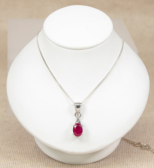 A silver chain necklace with an oval-cut red ruby gemstone pendant, displayed on a white jewelry bust against a neutral beige background.
